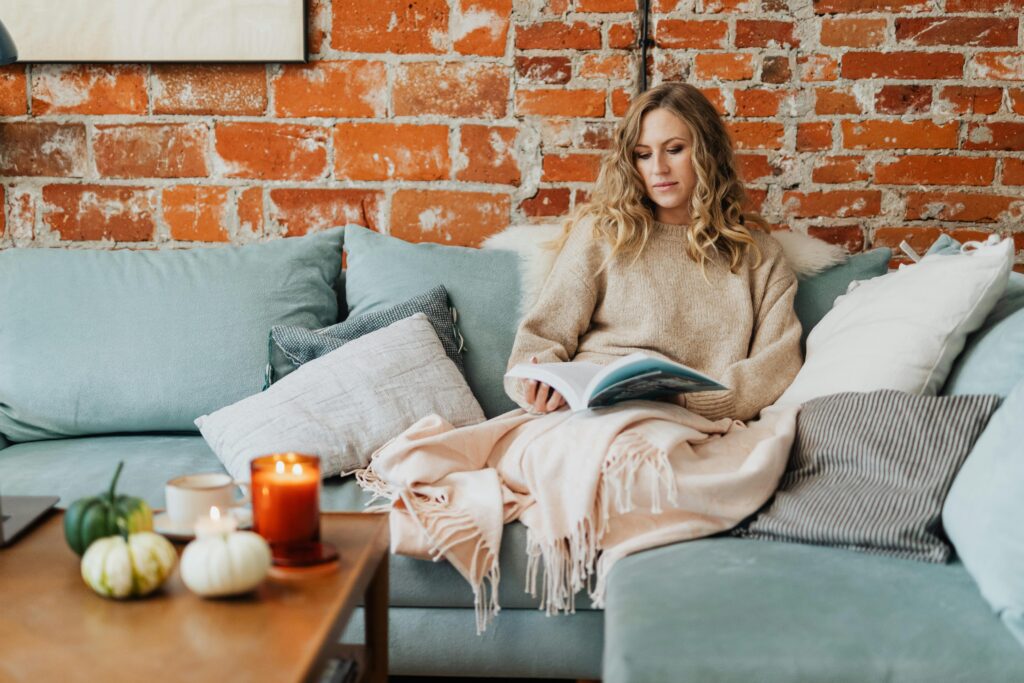 A woman enjoys a peaceful reading moment on a comfy sofa with lighted candles and a warm, cozy ambiance.