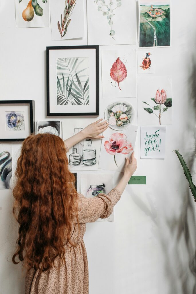 Woman arranging a collection of framed botanical prints on a wall, showcasing her artistic taste.