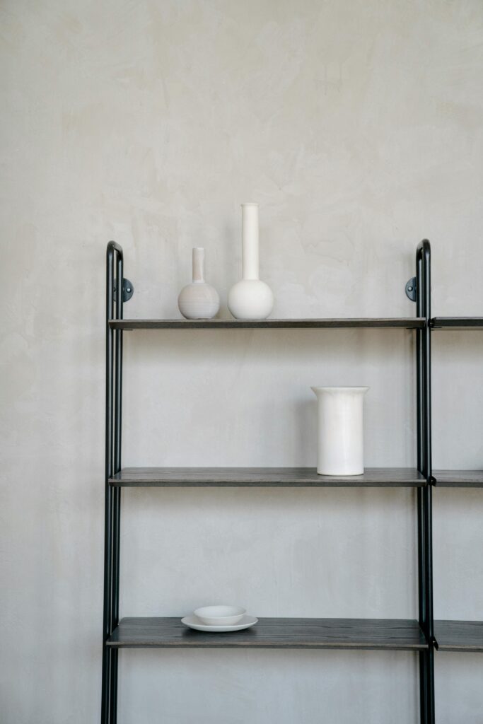 A minimalist black metal bookshelf with white ceramic vases on a neutral wall.