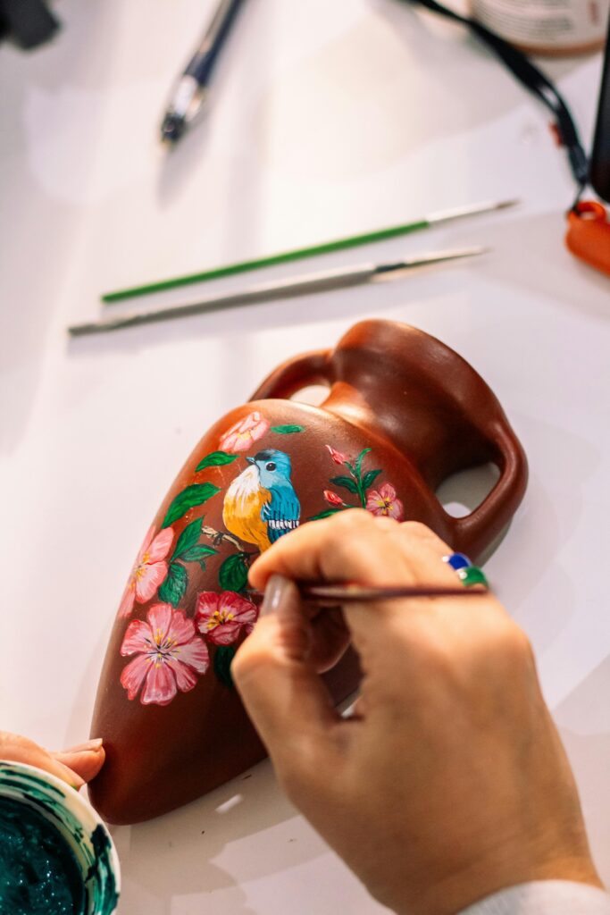 Artisan hand-paints vibrant floral and parrot design on pottery vase. Perfect for art and craft themes.